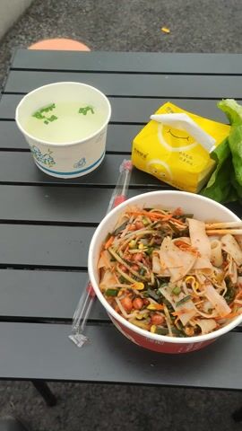 A top-down shot shows a meal on a dark grey slatted outdoor table. A white bowl filled with noodles, bean sprouts, peanuts, and leafy green vegetables is in the center. Two wooden chopsticks are resting in the bowl. To the left of the bowl, a yellow paper napkin dispenser with a smiley face design is partially obscured by a bunch of fresh green lettuce leaves. In front of the napkin dispenser, a small white paper cup with a blue rim contains a light-colored liquid with chopped green onions. A clear plastic straw is lying horizontally across the table, partially under the bowl. In the background, a terracotta pot with a white object on top is visible, and to the far right, another terracotta pot base is present. The ground beneath the table appears to be dark asphalt. A person's feet wearing black slide sandals are visible on the far left, walking away from the table.