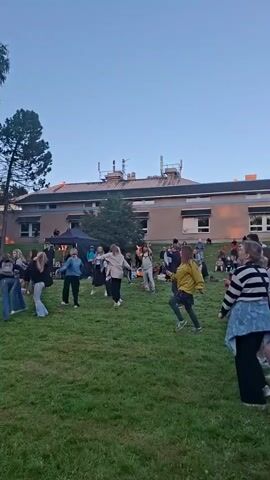 The video captures an outdoor gathering on a grassy lawn in front of a building. A group of people, of various ages, are spread across the lawn, some dancing. The setting appears to be in the late afternoon or early evening, with the sky a soft blue and the sun casting a warm glow on the trees. A makeshift stage is set up on the left side of the frame, with lights visible. A black tent is positioned further back on the right. The people are casually dressed, suggesting a relaxed atmosphere. The overall scene is one of community and celebration, possibly a concert or outdoor event.