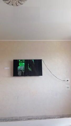 A flat-screen television is mounted on a textured, light-colored wall. The television is displaying a dark image with green elements, possibly a nature scene or a video game. A black cable runs from the television to two electrical outlets on the right side of the wall. Above the television, a white ceiling fixture with multiple lights is visible. The wall has a subtle, repeating pattern. The bottom of the image shows the top of a white baseboard. The scene is static, with no visible movement.