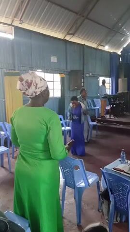 The video shows an indoor gathering, possibly a church service. In the foreground, a woman in a green dress faces away from the camera, her head covered with a floral scarf. She is surrounded by blue plastic chairs. In the background, several people are visible. Two women in dresses, one blue and one gray, appear to be talking or interacting. A man in a white shirt and dark pants is also in the background, facing away from the camera and facing the stage area. The environment appears to be a room with a corrugated metal ceiling and blue and white draped fabric covering the walls or backdrop of a stage. Windows are visible on the left side of the room. The lighting is somewhat dim. Some tables with papers or books are also present.
