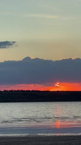 The video captures a serene sunset over a body of water. The sky is painted with hues of orange and pink, transitioning to a lighter blue towards the top. Dark, thick clouds partially obscure the setting sun, creating a dramatic effect with streaks of bright orange light peeking through. Below the sky is a dark, silhouetted line of vegetation or trees along the horizon. The water in the foreground reflects the vibrant colors of the sunset, with gentle ripples disturbing the surface. The immediate foreground appears to be a sandy or muddy shoreline. The overall atmosphere is peaceful and tranquil. There are no people or distinct objects visible besides the natural landscape.