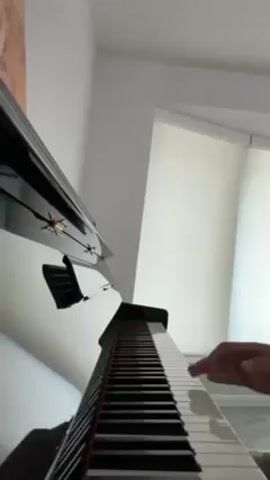 A low-angle shot shows a pair of hands playing a glossy black piano. The piano's keyboard fills the bottom half of the frame. Above it, the piano's open lid reflects the room, and a music stand is visible. To the right is a window with white blinds, and a white wall is in the upper left. The hands are fair-skinned, with the left hand casting a shadow on the piano's surface. The hands play the keys, and a bracelet is visible on the left wrist.