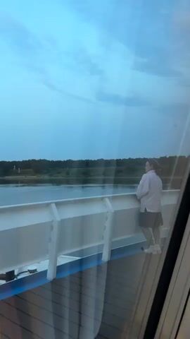 The video shows a woman standing on the deck of a ship, looking out at a body of water. The sky is a pale blue, and there are some clouds. The water is calm, and there is a line of trees in the distance. The woman is wearing a light-colored shirt and a skirt. She is standing with her hands clasped in front of her. The camera is positioned inside the ship, looking out through a window.