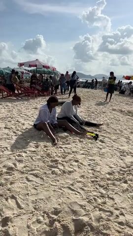 Two women sit on a sandy beach. The woman on the left wears a white shirt and black shorts. The woman on the right wears a white top and black leggings. Both women are facing towards the right side of the frame. In front of the woman on the right, there is a snorkel mask and a yellow snorkel. There are other people on the beach behind them, some standing and some sitting. There are beach umbrellas in the background, and mountains are visible in the distance. The sky is cloudy.