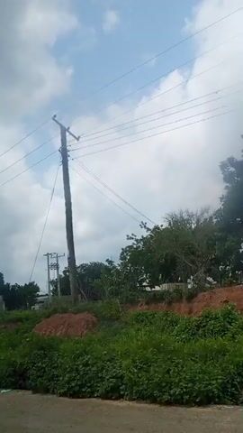 The video begins with a wide shot of a billboard featuring a man in a suit with a green logo at the top. The billboard is situated on a grassy incline beside a road. The camera pans to the right, showing a partially constructed building with cinder block walls, some construction materials, and greenery. Further to the right, a large tree stump is visible on the side of the road, along with more construction materials. The video then shows a person riding a motorcycle on the road, passing by trees and power lines. Finally, the camera pans to the right again, revealing a pile of large sacks on the side of the road, surrounded by trees and under a cloudy sky.