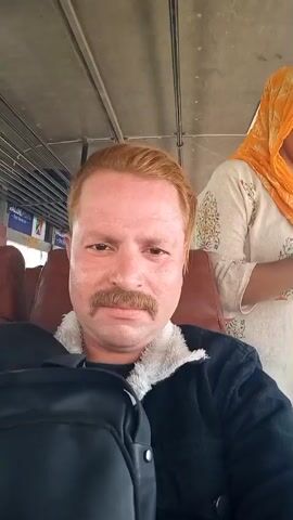 The video shows a man with reddish-blond hair and a mustache, sitting on a bus, wearing a black jacket with a white, furry collar. He is looking directly at the camera for most of the video. At one point, the camera swings to the side, and then back. Behind him, a woman wearing an orange headscarf and patterned white shirt is standing. The bus has brown seats and metal bars. The video appears to be taken from a passenger's perspective.
