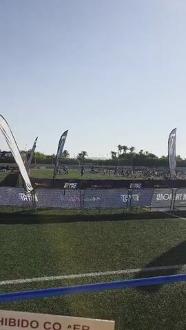The video pans across a grassy field where a triathlon event is likely taking place. In the foreground, there is a silver metal fence with banners attached. The banners display event logos and text, including "MEDITERRANEA TRI" and hashtags. Beyond the fence, the field is covered in numerous bicycles parked in rows, suggesting a transition area or race staging. Several people are visible, some standing near the bicycles, while others are moving around the area. A few individuals appear to be riding bicycles in the background. Tall, thin flags with event branding are positioned at intervals. Palm trees and a few buildings or structures are visible in the background, under a clear blue sky. The overall scene depicts an outdoor sporting event in a sunny environment.