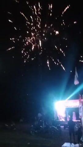 The video captures a night scene with fireworks exploding in the dark sky. Below, a stage is illuminated by bright spotlights, some of which project upwards. Several people are visible in the foreground, appearing to watch the fireworks or the event on stage. Motorcycles are parked in the area. A flag is seen flying from a pole above the stage. The overall atmosphere suggests a celebration or outdoor event.