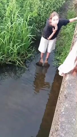 The video shows two people standing on the edge of a concrete structure that overlooks a body of murky water with tall green grass on the opposite bank. One person, a young individual wearing a black hoodie and light-colored shorts, is standing in the water up to their knees. This person is facing the water and appears to be reaching down. Their reflection is visible in the water. Another person, partially visible on the right side of the frame, is wearing a white garment and is leaning over the edge, also looking at the water. The lighting suggests it is daytime.