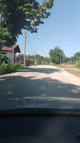 A car drives down a straight, paved road on a sunny day. Houses are visible on either side of the road, and there are telephone poles with wires running alongside the road. There are trees that create shadows on the road. A blue road sign is visible in the distance. The sky is clear and blue.