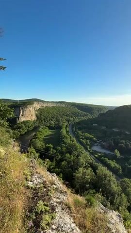 The video shows a panoramic view of a lush green valley with a river winding through it. The valley is surrounded by steep, tree-covered hills. A road and a railway track run along the river. There are a few buildings scattered in the valley. The sky is clear and blue, with the sun shining brightly. The perspective is from a high vantage point, looking down into the valley. The foreground is dominated by rocky terrain with sparse vegetation. The overall impression is one of a serene and natural landscape.