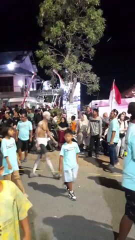 The video shows a nighttime street scene in what appears to be a public gathering or celebration in Kecamatan Tanggulangin, Indonesia. Many people of various ages, including adults and children, are present. Several children are wearing matching light blue t-shirts. A distinctive figure, a shirtless man covered in white body paint or powder and wearing only dark shorts, walks through the crowd. An Indonesian flag (red and white) is visible in the background, suggesting a patriotic or cultural event. The setting is illuminated by artificial lights, casting shadows on the ground. A large tree stands prominently in the foreground. People are standing, walking, and observing, indicating a festive or communal atmosphere.
