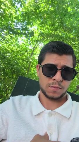 A medium shot shows a man in sunglasses sitting in a chair. He has dark hair and a trimmed beard, and he is wearing a white polo shirt. The camera angle is low, looking slightly up at him. Behind him is a backdrop of green leaves and tree branches, dappled with sunlight. The man adjusts his shirt at the beginning of the video, then looks directly at the camera with a neutral expression.
