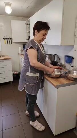 A middle-aged woman stands in a kitchen, wearing a striped apron, gray leggings, and light-colored sandals. She is working at a white countertop, mixing ingredients in a stainless steel bowl. To her right, another bowl sits on the counter, and behind her is a sink with a faucet. The kitchen has white cabinets, a tile floor, and a light fixture on the ceiling. A set of knives is mounted on the wall to her left, and a white dresser is visible in the background. She glances at the camera.