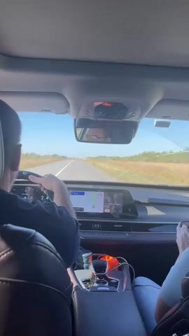The video shows the interior of a car from the perspective of a rear passenger, looking forward. A driver's hands are visible on the steering wheel, and the driver's head and upper body are seen in the rearview mirror. The car is on a paved road with a clear blue sky and dry, grassy fields on either side. The dashboard features a large display screen showing navigation. A second person's legs and hands are visible in the lower right of the frame, holding a phone. The overall scene depicts a daytime car journey through a rural landscape.