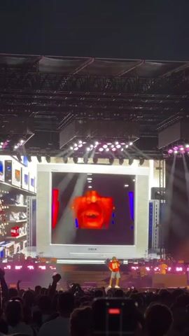 A wide shot captures a live concert performance. A large screen dominates the stage, displaying a green screen with silhouetted figures dancing. In front of the screen, a man in a red shirt and shorts stands on a platform, facing the audience. He appears to be performing or interacting with the visuals. The stage is lit with purple and white lights. A crowd of people is visible in the foreground, many holding up their phones to record the event. The overall environment appears to be an outdoor evening concert.