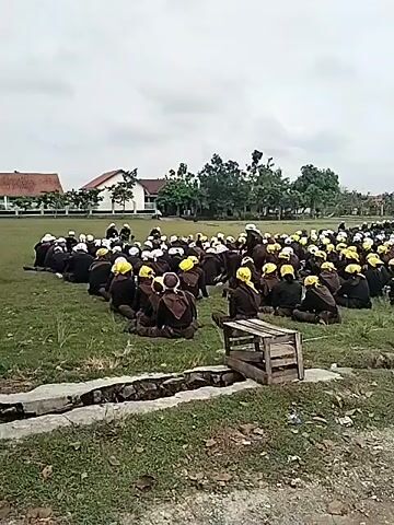 A wide shot captures a large group of individuals, predominantly male, gathered in an open grassy field under an overcast sky. The subjects are seated on the ground in organized rows, mostly facing away from the camera, towards an unseen point in the distance. They are dressed in dark-colored uniforms or clothing, with many wearing distinctive yellow or white hats or helmets.

In the foreground, a concrete path or road runs across the bottom of the frame, with a ditch or drainage canal separating it from the grassy area where the people are seated. A rustic wooden crate or structure lies near the ditch. Briefly, a motorcycle is visible parked on the concrete path on the right side of the frame.

The background features several single-story buildings with red or dark-colored roofs, interspersed with numerous trees, suggesting a suburban or institutional setting. Some individuals appear to be standing further back in the field, possibly overseeing the seated group.

The overall scene suggests an outdoor assembly, training session, or briefing for a large organized group, possibly a cadet corps, community service group, or similar organization, given their uniform appearance and structured seating. The sky is uniformly cloudy, indicating an overcast day. A loud engine noise is audible at the beginning of the video, quickly fading. No specific text is visible in the frame. The location is Tawangsari, Indonesia.