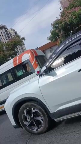 The video pans across a street scene with several vehicles. In the foreground, the right side of a white SUV is visible, showing its front wheel and headlight assembly. To the left of the SUV, a white minivan with orange accents on its side and rear is seen. Further down the street, another white car is parked, its license plate partially obscured. In the background, there are buildings, trees, and a street light. The sky is visible, with the sun shining. There are some shops on the side of the road, with signs visible. The overall environment appears to be an urban street.