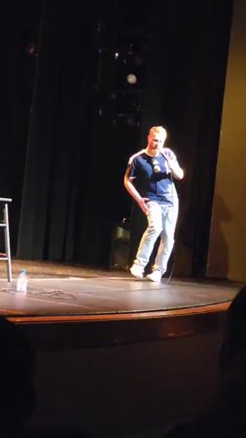 A man with red hair and a beard, wearing a blue t-shirt with a white and yellow emblem and light blue jeans, stands on a stage in front of a black curtain. He is holding a microphone in his right hand and gesturing with his left hand. To his left is a wooden stool with a bottle of water and what appears to be a cell phone on it. The stage is dimly lit, with spotlights visible in the background. The audience is not clearly visible, but silhouettes of people can be seen in the foreground. The man appears to be a comedian or performer, engaging with the audience.