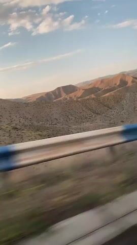 The video shows a fast-moving view from a vehicle, likely a car, looking out over a mountainous, arid landscape during the daytime. In the foreground, a blurred guardrail with blue and white markings runs horizontally across the bottom of the frame, indicating motion. The middle ground features rolling hills covered in sparse vegetation, appearing dry and desert-like. The background is dominated by more distant mountains with layered geological formations, bathed in the warm light of what appears to be sunset or sunrise. The sky above is blue with scattered white clouds, suggesting fair weather. There are no people or specific objects identifiable in the scene besides the guardrail. The overall impression is of travel through a remote, natural environment.