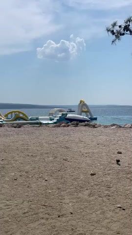The media file shows a sunny day at a beach in Crikvenica, Croatia. In the foreground, there is a sandy beach with scattered pebbles. In the background, a large inflatable water park is set up in the clear blue water. The water park features slides, climbing structures, and other inflatable obstacles. The sky is blue with a few fluffy white clouds. The sun is shining, creating a bright and cheerful atmosphere. The scene is likely set in the late morning or early afternoon, given the brightness of the sunlight. There are no people clearly visible in the frame, but the presence of the water park suggests a recreational area frequented by visitors. The calm water and clear skies indicate pleasant weather, ideal for beach activities. A small boat is visible in the water near the inflatable park. The overall impression is one of a relaxing and fun summer day at the seaside.