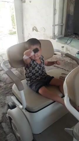 A young child sits on a white golf cart. The child is wearing a patterned tank top and gray shorts. The child is moving their arms and head. The background appears to be an outdoor area with a concrete floor and a white wall.
