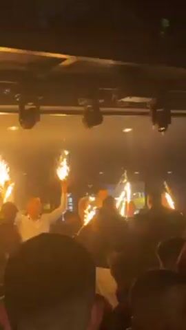 The video shows a crowded indoor venue, likely a nightclub or upscale bar. Several individuals, possibly staff, are holding sparklers aloft. The crowd appears to be celebrating, though their specific interactions are obscured by the density of people. The environment includes lighting fixtures, with beams of colored light intermittently illuminating the scene. The overall atmosphere suggests a festive and lively event.