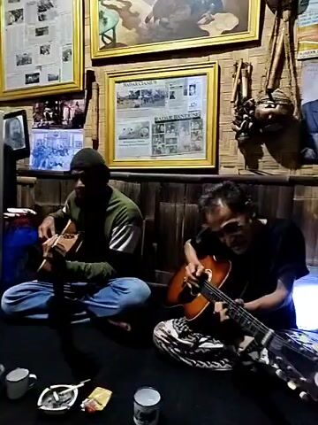 The video shows three people playing musical instruments in a room decorated with pictures and various objects. Initially, two men are sitting on the floor, one playing a small guitar and the other playing a stringed instrument that appears to be a lute. There are mugs, an ashtray, and a pack of cigarettes on the floor in front of them. The walls are adorned with framed pictures and other decorative items, suggesting a space with cultural or artistic significance. The camera then pans to reveal a third man seated on the floor in another part of the room. He is playing a bamboo instrument. The space around him seems cluttered, with various items scattered around, including bags, tools, and containers. There is a glass display case filled with objects and a window letting in natural light. The setting suggests a workshop or a cultural center where music is being created and preserved.