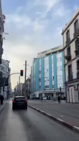 The video shows a city street on a cloudy day. A car waits at a red light on the left side of the street. In the center of the frame, tram tracks run down the middle of the street. Several pedestrians walk along the sidewalk on the right side of the street near a building called "Hotel Tandja". Buildings line both sides of the street. The street is wet, likely from recent rain. A bird flies across the sky.