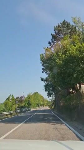 The video shows a view from the driver's seat of a car traveling on a paved road. The road is bordered by trees and vegetation on both sides, with a guardrail on the left. The sky is clear and blue. There are a few traffic signs visible along the road. The car is moving forward, and the scenery is passing by. The time stamps indicate the video is approximately 5 seconds long.