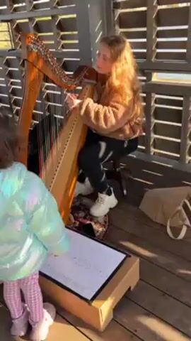 A young, light-skinned woman with long, auburn hair is sitting on a wheeled stool and playing a wooden harp. She wears a brown, fuzzy jacket, black leggings, and white sneakers.  She is sitting on a colorful rug. In front of her, resting on a cardboard box, is a white board with numbers and letters written on it. A little girl in a shiny, light blue jacket and pink plaid pants is standing near the box and looking at the woman. There is a beige tote bag next to the cardboard box. The scene is taking place in a structure with an open-slatted roof and walls, allowing sunlight to filter through.