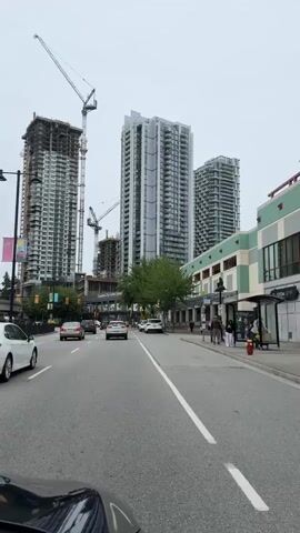 The video shows a street scene with multiple cars driving on the road, which has lane markings. On the right side of the street, there is a sidewalk with a few people walking. The buildings lining the street are tall and modern, with some under construction, indicated by cranes. The sky is overcast. The camera appears to be mounted inside a moving vehicle.