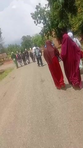 A group of people are walking down a paved road. In the foreground, two women in hijabs, one red and one maroon, walk away from the camera. Behind them, a group of individuals, some in what appear to be military or security uniforms, follow. Further in the background, a building with multiple windows is visible, surrounded by trees and dry vegetation. The sky is overcast.