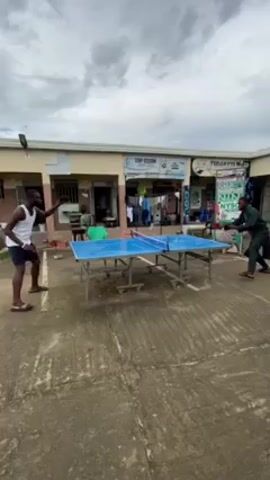 In a medium shot captured outdoors under a cloudy sky, two men are engrossed in a game of ping pong. The man on the left is attired in a white tank top and dark shorts, while the man on the right sports a dark green suit. They are both in motion, actively engaged in hitting the ping pong ball across a blue ping pong table with a net in the middle. In the background, a row of single-story buildings is visible, interspersed with doorways and signs for businesses such as "TOP CLEAN" and "IBADAN". The paved ground surrounding the table is aged and weathered. Several other individuals are present in the background, though their activities are obscured by distance and the primary focus on the ping pong match.