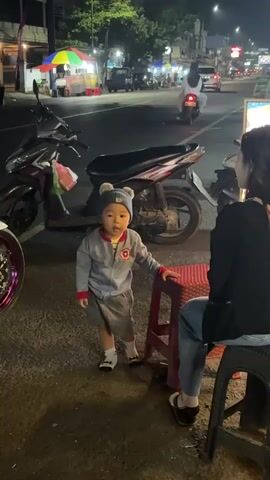 A young child wearing a grey jacket, grey shorts, a grey hat with pom-poms, and white socks stands on a sidewalk at night next to a red plastic stool. The child is looking at something to the right. A woman wearing a black top and blue jeans is sitting on a black plastic stool to the right of the child, with her back mostly to the camera. In the background, a street is visible with several motorcycles parked along the side. There are also streetlights, shop fronts, and vehicles moving on the street. The scene appears to be an outdoor food stall or market area.