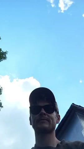 A person wearing a brown t-shirt, a brown baseball cap, and dark sunglasses is filmed from the chest up, looking at the camera. The person is outdoors with a blue sky and white clouds in the background. A house with a light blue exterior is visible to the right, and lush green foliage surrounds the person. At 00:02, the person raises their left hand and waves. The video also features a brief shot looking up at the sky, with trees visible in the upper left corner and the person's head and shoulders silhouetted against the bright sky.