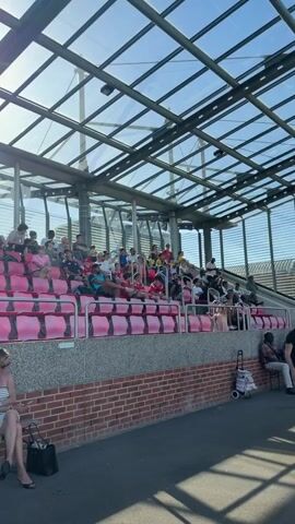The video shows a small crowd seated in pink stadium-style seats under a glass roof. People of various ages are seated and cheering. There is a woman in a striped dress sitting on a brick bench in the foreground. The seats are arranged in sections labeled "Bloc A" and "Bloc B." The sun is shining brightly through the glass roof.
