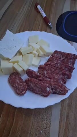 A close-up overhead shot displays a white plate of food on a wooden surface. The plate contains several types of cheese and sliced salami. To the left of the plate are two larger pieces of white cheese, one appearing crumbly and the other smoother. In the center, several cubes of yellow cheese are arranged. To the right, thin slices of red salami are neatly stacked. In front of the salami slices are two smaller, whole salami sausages. A red and white pen rests in the background, along with a black circular object. The lighting is soft, and the focus is on the food.