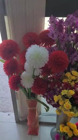 The video pans across a collection of artificial flowers arranged in vases and buckets. The flowers are of various colors, including red, white, yellow, pink, purple, and blue. Some of the flower arrangements are quite large and elaborate, while others are smaller and more delicate. In the background, there are two statues of women holding pots with greenery. The scene appears to be indoors, possibly a shop or a home, with natural light coming from a window. The overall impression is one of a vibrant and colorful display of floral decorations.