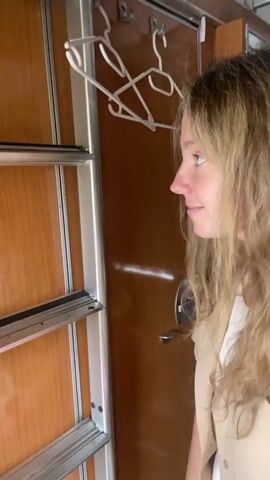 The video opens with a medium shot of a young woman with blonde hair, smiling at the camera. She is wearing a white top and a tan vest. Behind her, the interior of a train car is visible, with wooden paneling and metal shelving. The camera then pans to the left, showing the exterior of the train passing by. We see a brief glimpse of another passenger in the train car, and then the view shifts to the landscape outside. The scene outside is a mix of trees, buildings, and a street with a parked car. The sky is cloudy. The video appears to be a casual recording of a train journey.