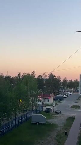 The video pans from right to left across a scene at dusk. In the foreground, there is a patch of green grass and a dark grey object that appears to be a covered trailer. A blue and black fence runs horizontally across the middle ground. Beyond the fence, there is a paved road with parked cars and a small building with a red roof and the letters "WD" visible on its sign. A line of trees, primarily birch with light bark, stretches across the landscape. In the background, more trees and buildings are visible, with a pale pink and orange sky above. On the far left of the frame, a multi-story building with a teal roof and brightly colored window frames is partially visible. The camera movement is smooth and steady.