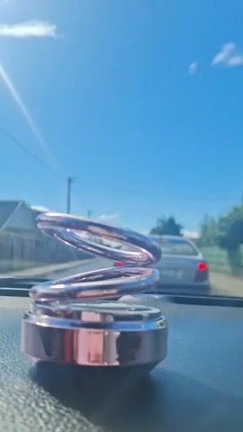 This media file captures a close-up view of a decorative car air freshener, likely placed on the dashboard. The air freshener is a metallic, spiraling sculpture with multiple rings, designed to catch light and potentially rotate. The background reveals a clear blue sky with scattered white clouds, suggesting a bright, sunny day. Sunlight glints off the air freshener and casts a lens flare effect in the upper left corner.

In the blurred background, a street scene is visible. A light-colored car with its brake lights illuminated is driving away, and houses and trees line the road. The overall setting appears to be a suburban or residential area during daylight hours, possibly mid-morning or early afternoon given the brightness and angle of the sun. There is no discernible text within the media file. The focus is on the aesthetic appeal of the air freshener and its interaction with the natural light and movement of the car.