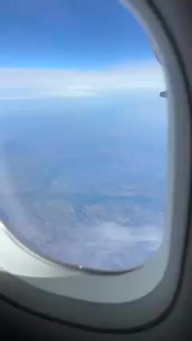 The video shows a view from an airplane window, looking down at the land below. The sky above is bright blue. The land has a patchwork appearance. Wisps of clouds are visible both near the top of the frame and closer to the ground.
