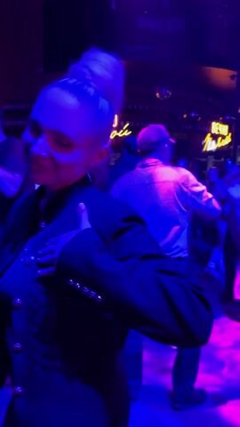 The video shows a group of people dancing in a dimly lit nightclub with a blue and purple lighting scheme.  The camera focuses on individuals as it moves through the crowd.  At first, a woman in a black jacket with gold buttons is seen dancing close to the camera. Then, the perspective shifts to the backs of people dancing, revealing more of the environment, including a neon sign in Cyrillic letters, which reads "ВЕЧНО Молодой". The background reveals bottles on shelves, suggesting a bar area.  The people are dressed in a variety of clothing. The atmosphere is lively, filled with movement and social interaction.