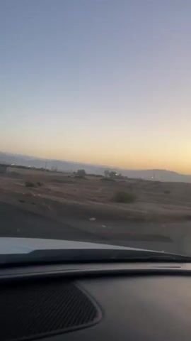 The video is a first-person perspective from inside a vehicle, looking out at a desert landscape during what appears to be sunrise or sunset. The sky transitions from a pale blue at the top to a soft orange and yellow near the horizon. The landscape is predominantly dry, brown earth with sparse, scrubby vegetation. In the distance, there are low hills or mountains, and a faint line of trees can be seen. There are also utility poles with wires stretching across the scene. The vehicle's dashboard is partially visible in the foreground, with a black, textured surface and a white edge. The video is slightly shaky, indicating movement of the vehicle.