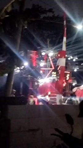 The video captures a nighttime outdoor event with a stage set up. The stage is adorned with numerous lights, mostly red, and several speakers. A tall, red and white striped pole stands prominently in front of the stage. In the foreground, a motorcycle is parked, with a wall partially obscuring the view. Several people are visible in the background, appearing to be attendees or participants, some of whom are silhouetted against the bright stage lights. The overall atmosphere suggests a lively gathering or performance.