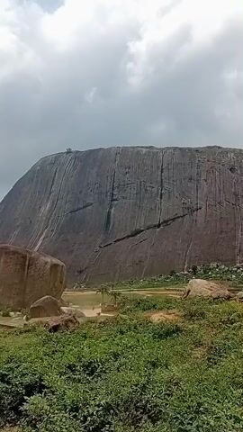 This is a landscape video showing large rock formations and vegetation under a cloudy sky. The video begins with a close-up of a massive, sheer rock face with visible vertical lines. To the left, several large boulders are scattered across the landscape. In the middle ground, there appears to be a body of water and some scattered figures, possibly people, near the water's edge. The foreground is covered in green grass and smaller rocks. The camera pans slightly to the right, revealing more of the rocky terrain and vegetation. The sky is predominantly cloudy with patches of brighter sky visible. The overall impression is one of a natural, possibly remote, rocky landscape.