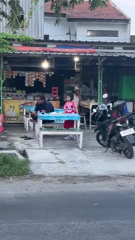 A man and a young girl are seated at a white picnic table outside a food stall. The man is wearing a blue shirt and dark pants, and the girl is wearing a pink shirt and red skirt. They are both looking down at the table. There is food on the table. Behind them, the food stall is filled with various packaged goods and merchandise. A motorbike is parked to the right of the table. The scene appears to be an outdoor dining area in front of a shop.