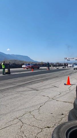 The video captures a drag race event set against a scenic backdrop of mountains under a clear blue sky. Two cars, a red coupe and a silver Volkswagen Golf, are positioned on a cracked asphalt track marked with black tire streaks. The race is lined with rows of spectators on both sides, secured behind tire barriers. Personnel in safety vests are stationed along the track, overseeing the event. As the race begins, both cars accelerate rapidly, emitting tire smoke. The Volkswagen Golf pulls ahead slightly, while the red coupe appears to drift slightly. The scene is captured from a ground-level perspective, offering a view of the crowd, the cars, and the surrounding landscape.