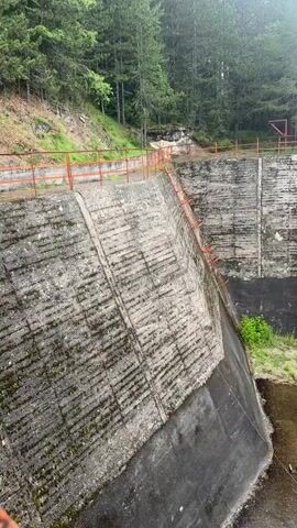 This media file shows a view of a concrete dam structure, likely a spillway or outlet works, in a forested area. The dam is made of rough, textured concrete with visible horizontal lines, possibly from the construction process. A red metal railing runs along the top edge of the dam, and a metal ladder is attached to the side of the dam, descending into the lower basin.

The basin below the dam is partially filled with murky water and has areas of dry concrete and vegetation. In the center of the basin is a circular concrete structure with a metal grate at the top, possibly a drain or an intake for a tunnel. There's another metal dome-shaped structure on the ground near the circular structure. The overall scene suggests an outdoor, possibly remote, location.

The lighting suggests it is daytime, and the overcast sky indicated by the diffused light and the green, lush vegetation imply a humid or potentially rainy weather condition. The environment is characterized by dense evergreen trees surrounding the dam. There is no visible text in the media file. No people are present, and no specific activity or event is clearly depicted, other than the structure itself and its immediate surroundings. The scene evokes a sense of disuse or perhaps maintenance in progress, given the exposed nature of the infrastructure.